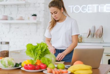 Woman in white t-shirt preparing fresh vegetables in a bright kitchen with a laptop.