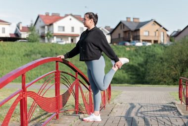 A woman in active wear performs a stretching exercise on a red bridge in a suburban park setting.